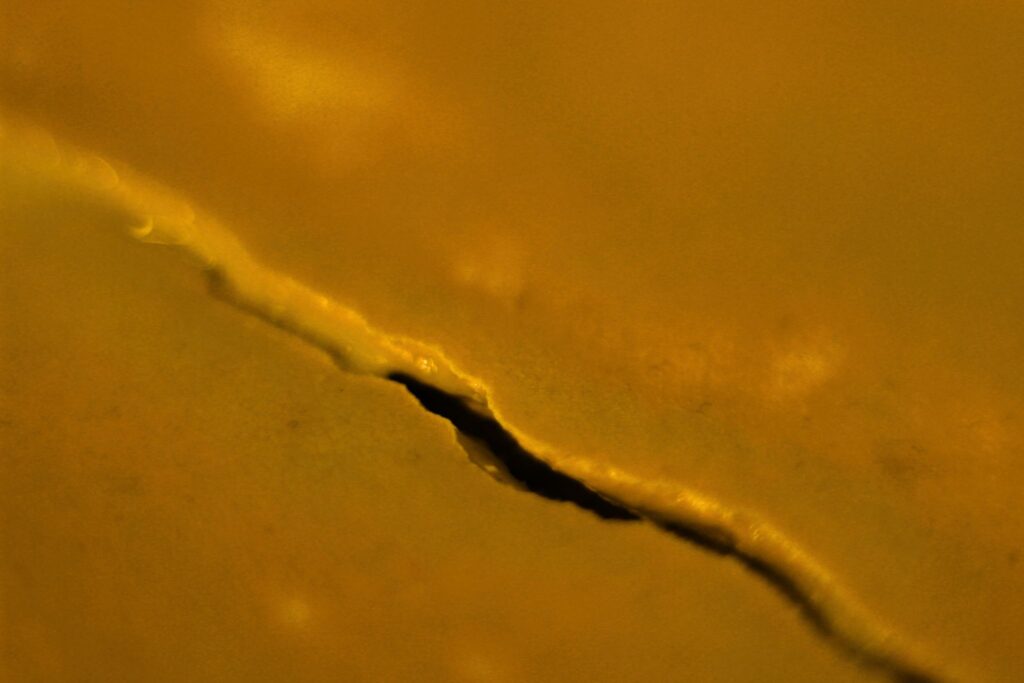 Abstrakte Makroaufnahme einer diagonal verlaufenden, dunklen Bruchlinie auf einer goldglänzenden Oberfläche mit weichen Lichtreflexen.