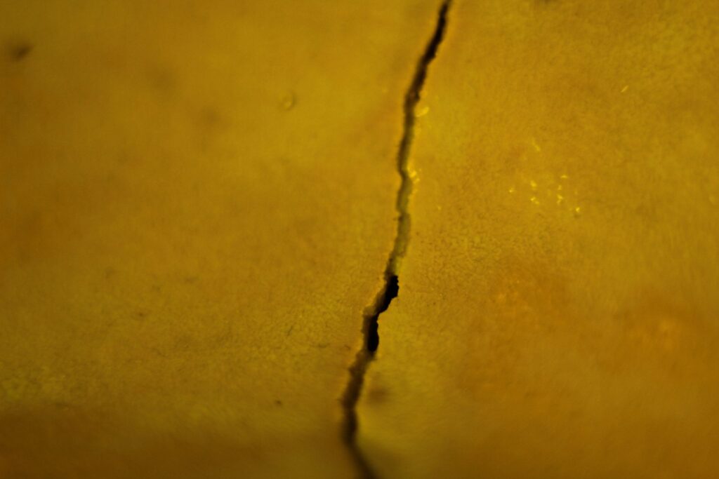 Abstrakte Detailaufnahme einer vertikalen, fein strukturierten Bruchlinie auf einer goldgelben Oberfläche mit weichem Schattenwurf.