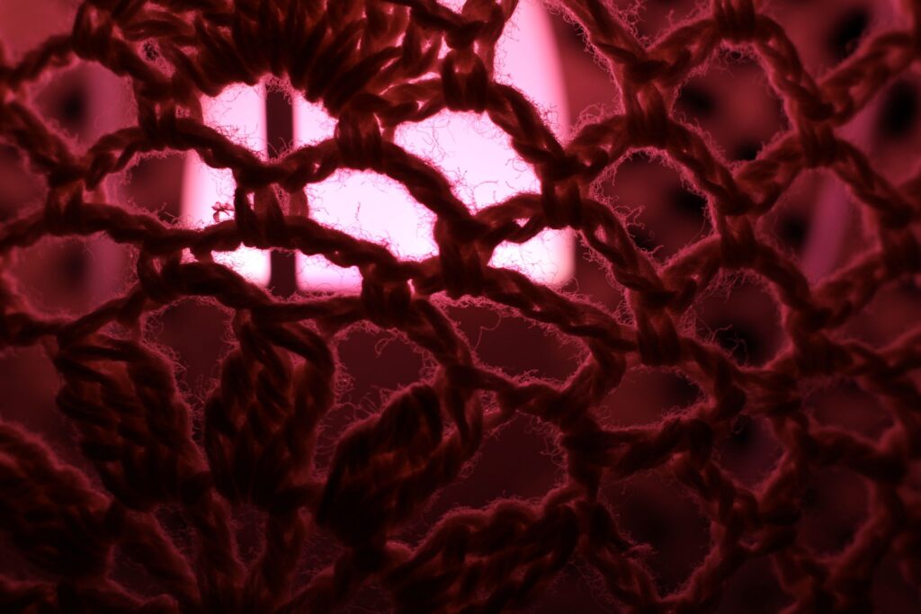 Abstrakte Makrofotografie eines dunklen Textilgewebes vor magentafarbenem Licht; künstlerische Detailaufnahme von Häkelstrukturen und feinen Fasern.