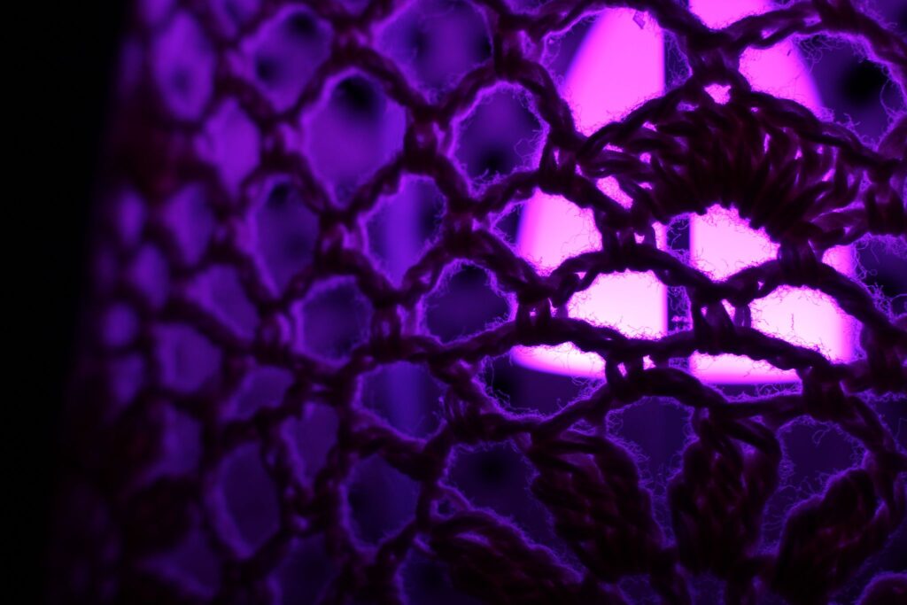 Abstrakte Makrofotografie einer Textilstruktur in intensivem violettem Licht; künstlerische Detailaufnahme von Häkelmaschen und Garnfasern mit leuchtendem Hintergrund.