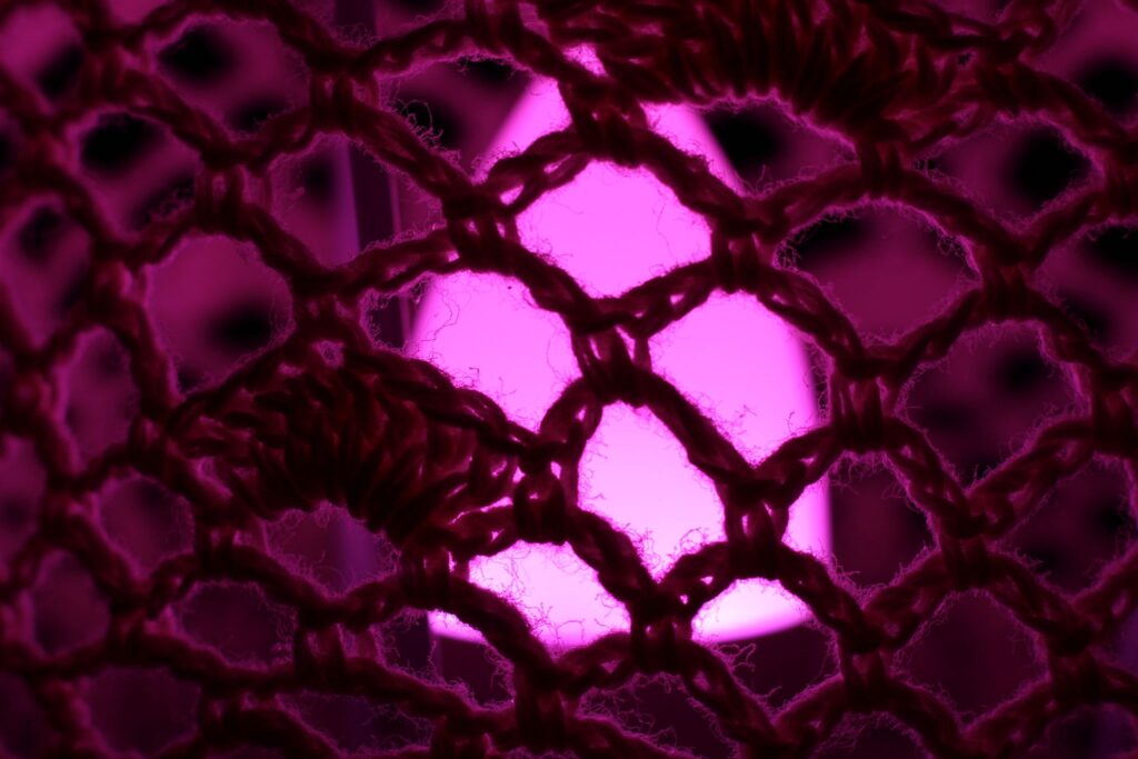 Abstrakte Makrofotografie eines Textilgeflechts vor leuchtend pinkem Hintergrund; künstlerische Detailaufnahme von Häkelstrukturen und feinen Garnfasern mit symmetrischem Lichtspiel.