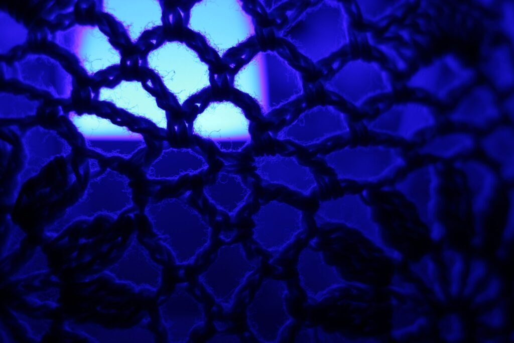 Abstrakte Makrofotografie eines dunklen Textilgewebes in tiefblauem Licht; künstlerische Detailaufnahme von Maschenstrukturen und Garnfasern vor leuchtend blauer Lichtquelle.