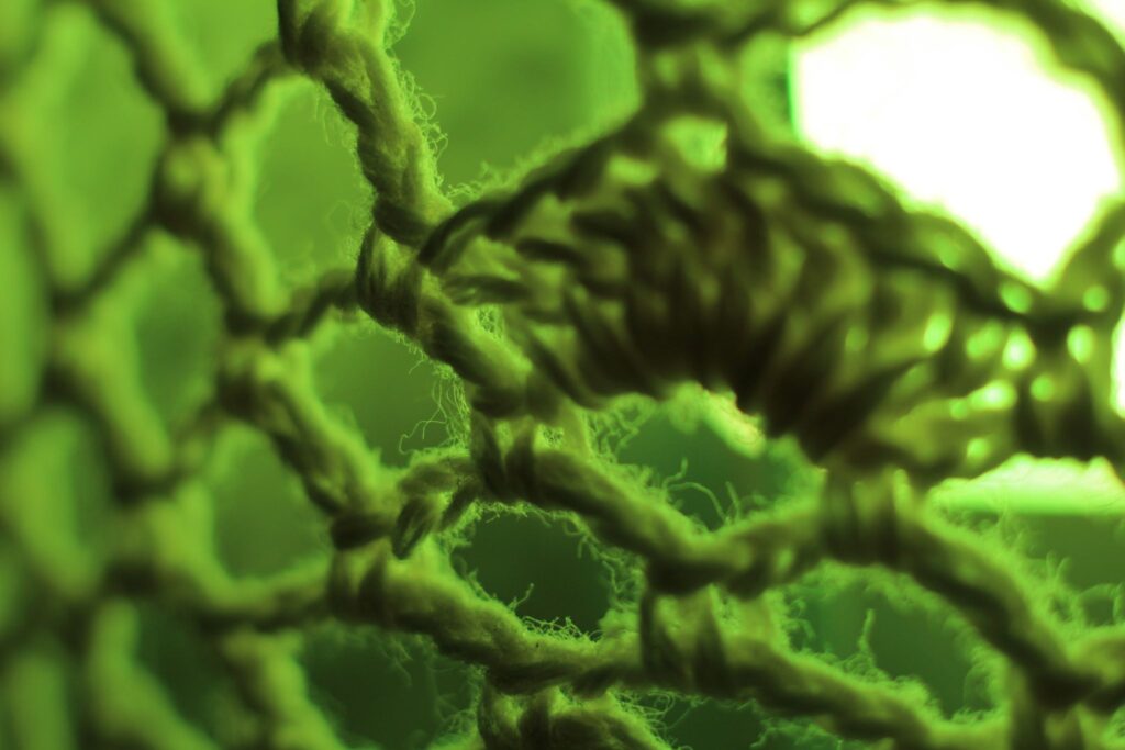 Abstrakte Makrofotografie eines Textilgeflechts in leuchtend grünem Licht; künstlerische Detailaufnahme von groben Garnschlingen und Fasern vor heller Lichtquelle.