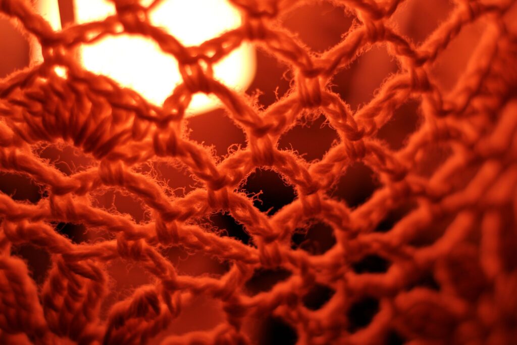 Abstrakte Makrofotografie eines Textilgeflechts in intensivem, glühendem Orange; künstlerische Detailaufnahme von Maschenstrukturen und feinen Garnfasern vor einer hellen Lichtquelle.