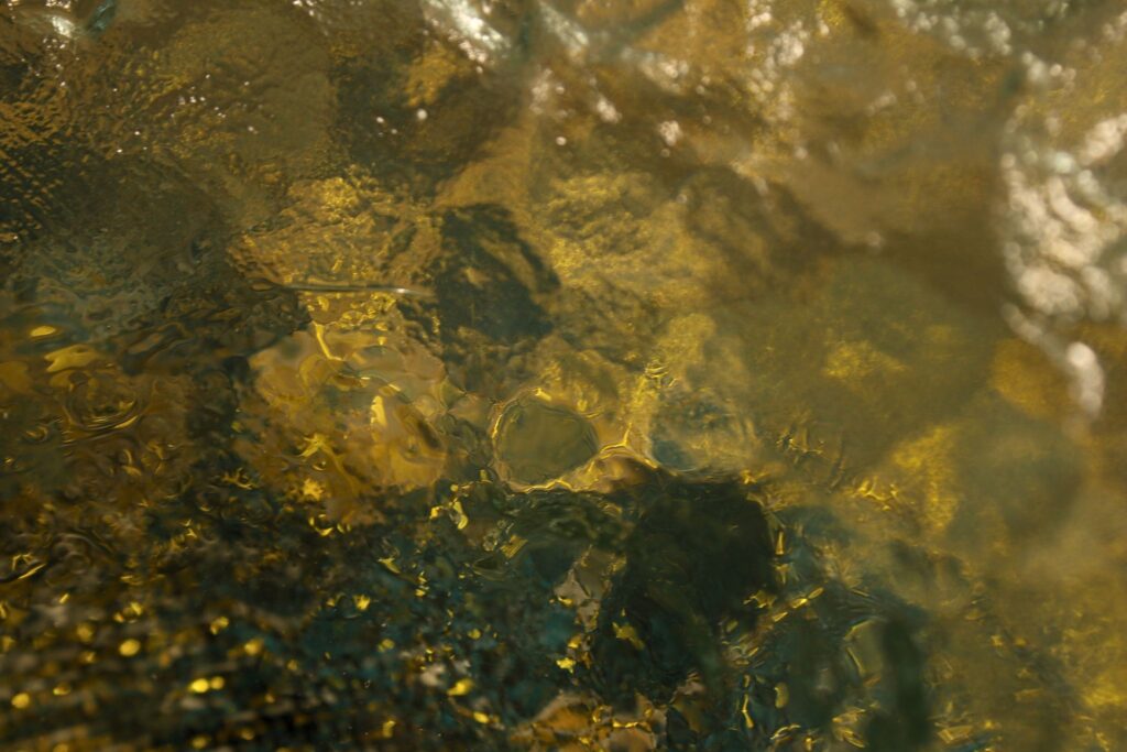 Ozean-Glas-01 Abstrakte Nahaufnahme einer wellenartigen Glas- oder Wasseroberfläche mit warmen Lichtreflexen in Goldtönen und dunklen Tiefen.